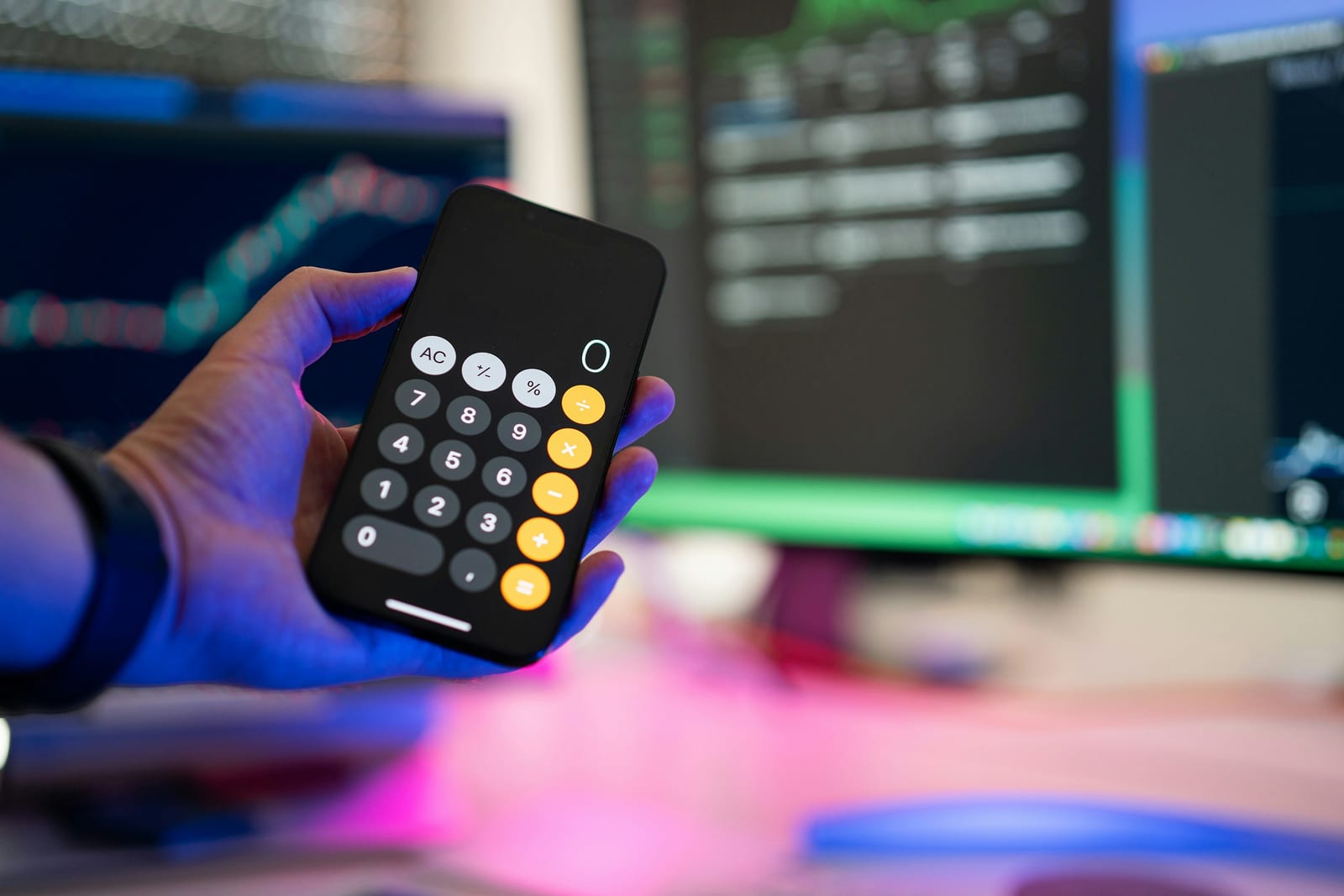 A hand holds a black calculator with illuminated buttons against a blurred background of multiple computer monitors…